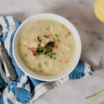 Creamy chicken and vegetable chowder served in a white bowl with green onions on top, placed on a rustic marble surface with a blue and white striped napkin, spoon, and condiments nearby.