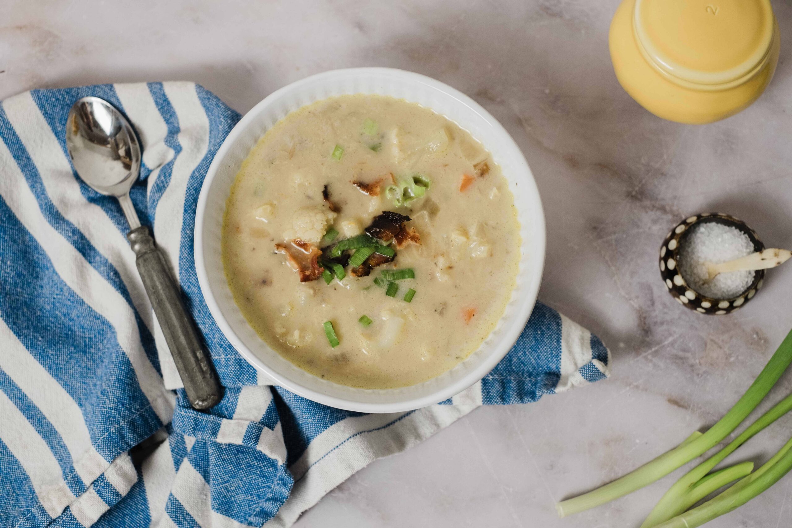 Creamy chicken and vegetable chowder served in a white bowl with green onions on top, placed on a rustic marble surface with a blue and white striped napkin, spoon, and condiments nearby.