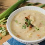Creamy homemade potato and vegetable soup garnished with chopped green onions in a white bowl on a rustic wooden table with fresh scallions and seasoning in the background.