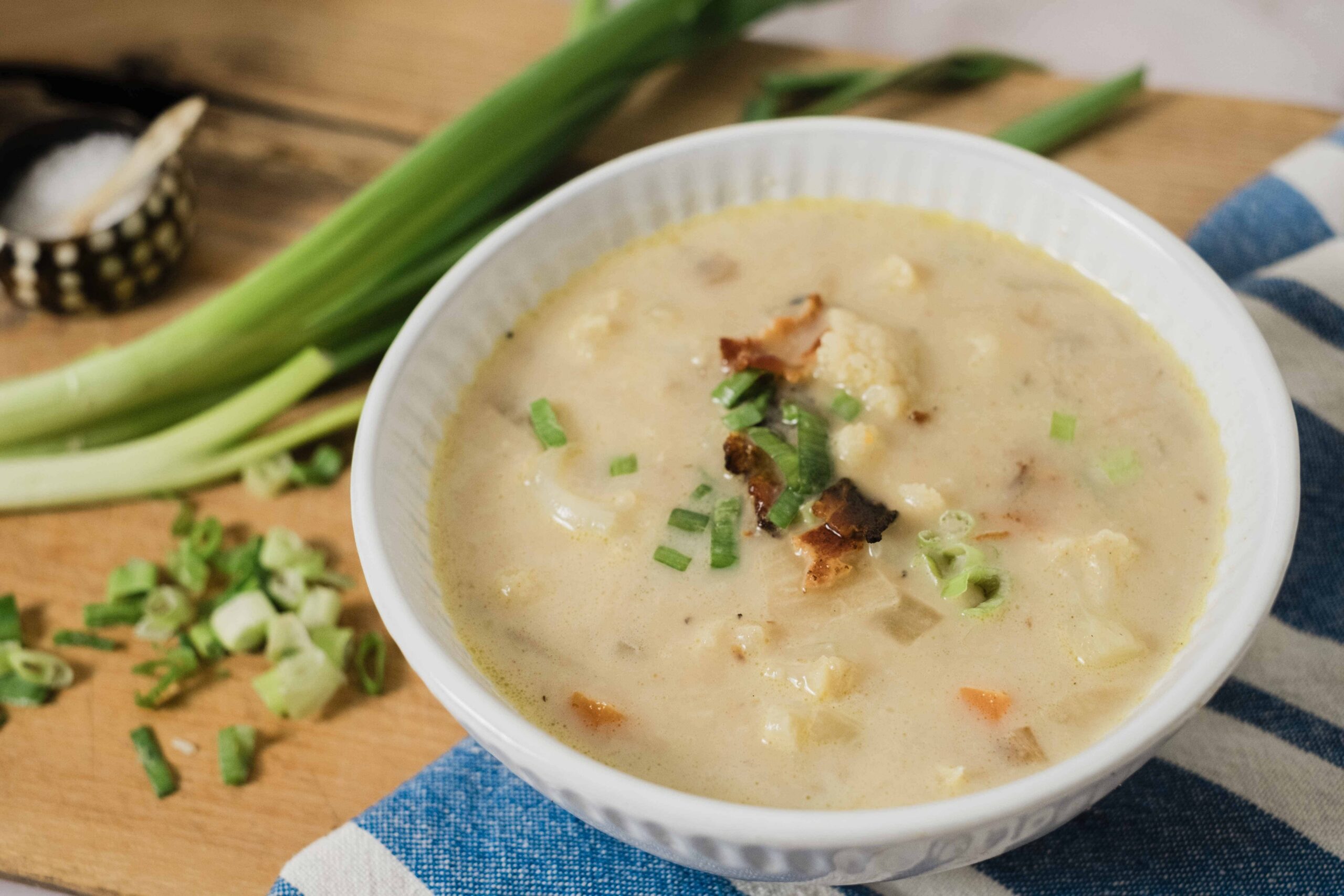 Creamy homemade potato and vegetable soup garnished with chopped green onions in a white bowl on a rustic wooden table with fresh scallions and seasoning in the background.