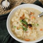 Creamy vegetable chicken pot pie soup with cauliflower, carrots, green onions, topped with crispy bacon bits and fresh herbs, served in a white bowl for a hearty homemade meal.