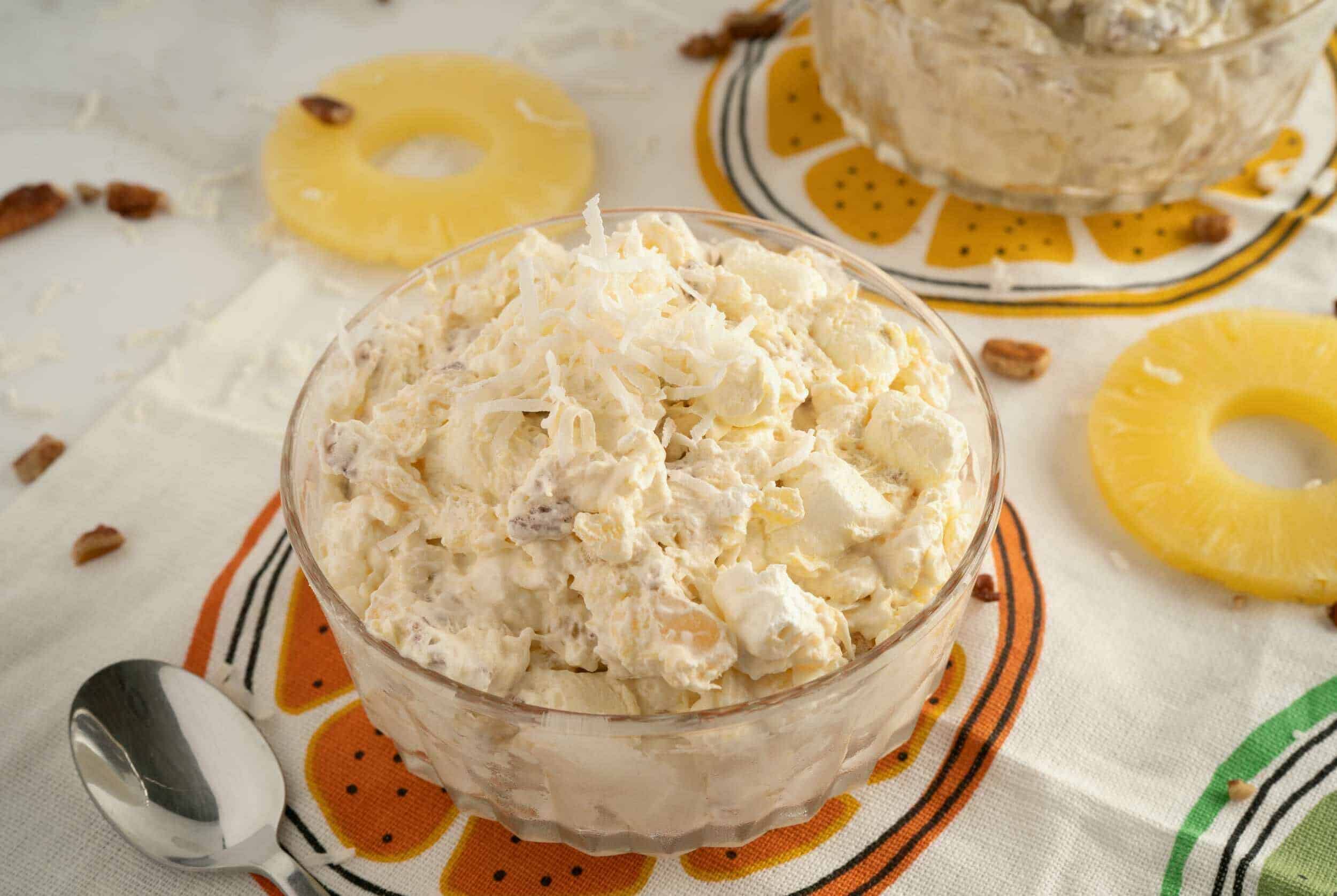 Creamy Hawaiian Macaroni Salad with pineapple chunks and chopped pecans in a glass bowl, served on colorful patterned tablecloth, fresh pineapple slices and scattered pecans for tropical flavor.