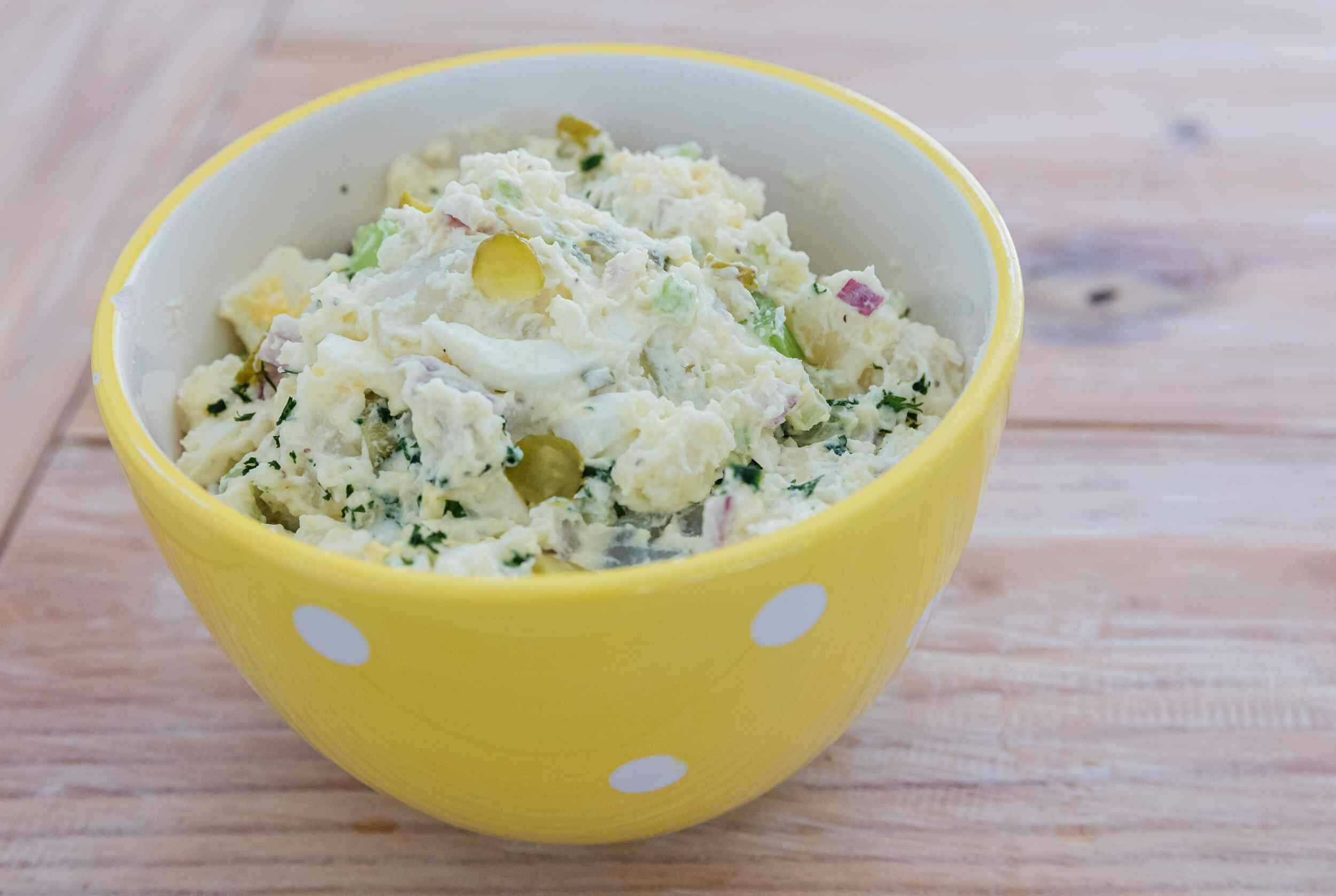 Creamy potato salad with chopped vegetables and herbs served in a yellow polka-dot bowl.