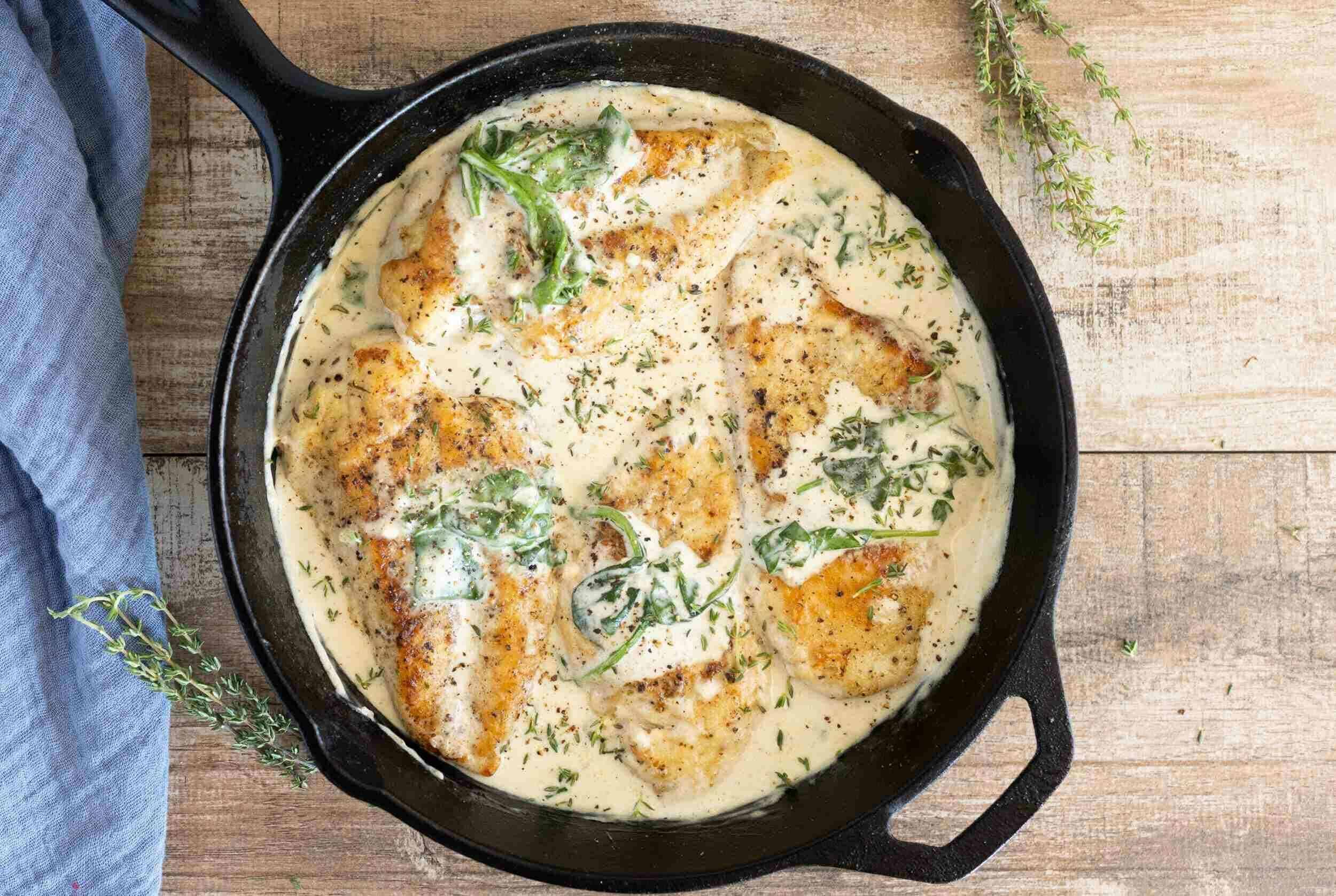 Juicy baked chicken breasts in a creamy herb sauce served in a black skillet on a rustic wooden table. Fresh thyme sprigs and a blue cloth are nearby, creating a warm, inviting meal presentation.