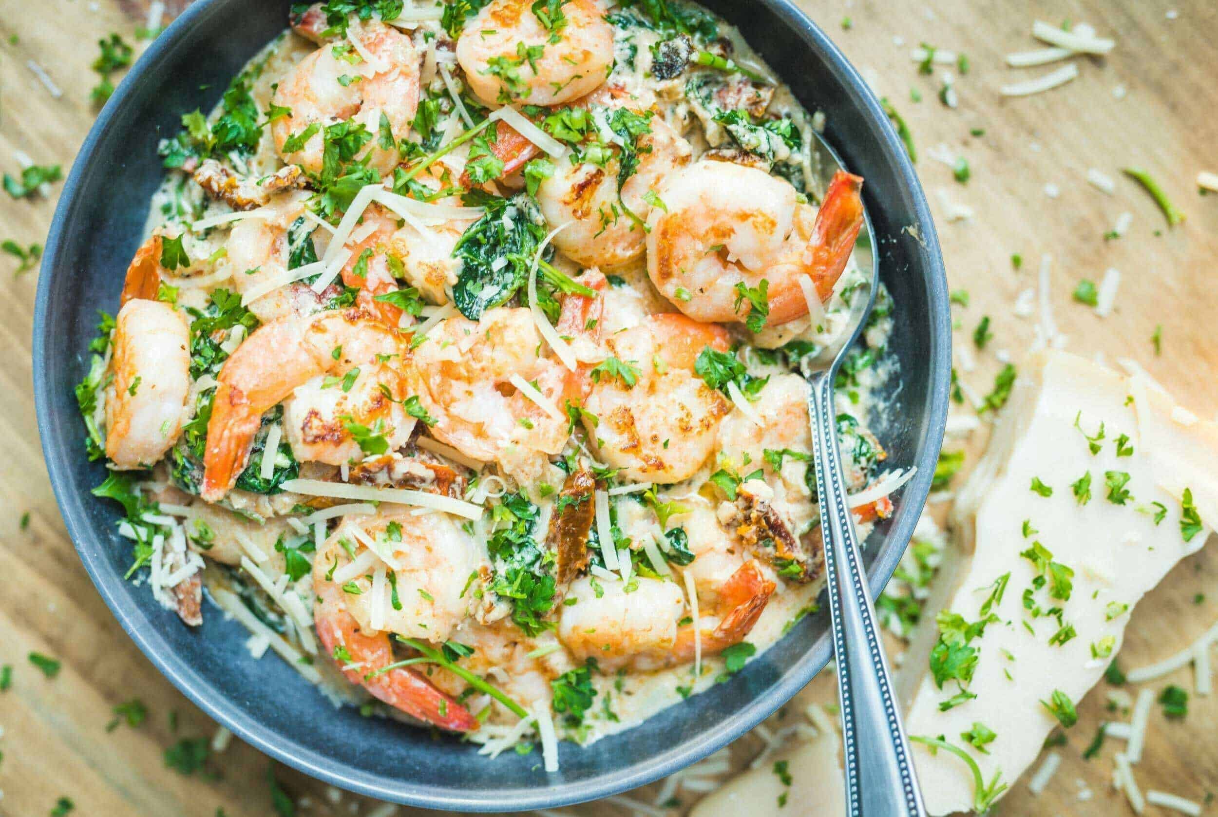 Juicy shrimp and fresh vegetables in a creamy pasta Alfredo dish topped with shredded cheese and chopped parsley, served in a black bowl on a wooden table.