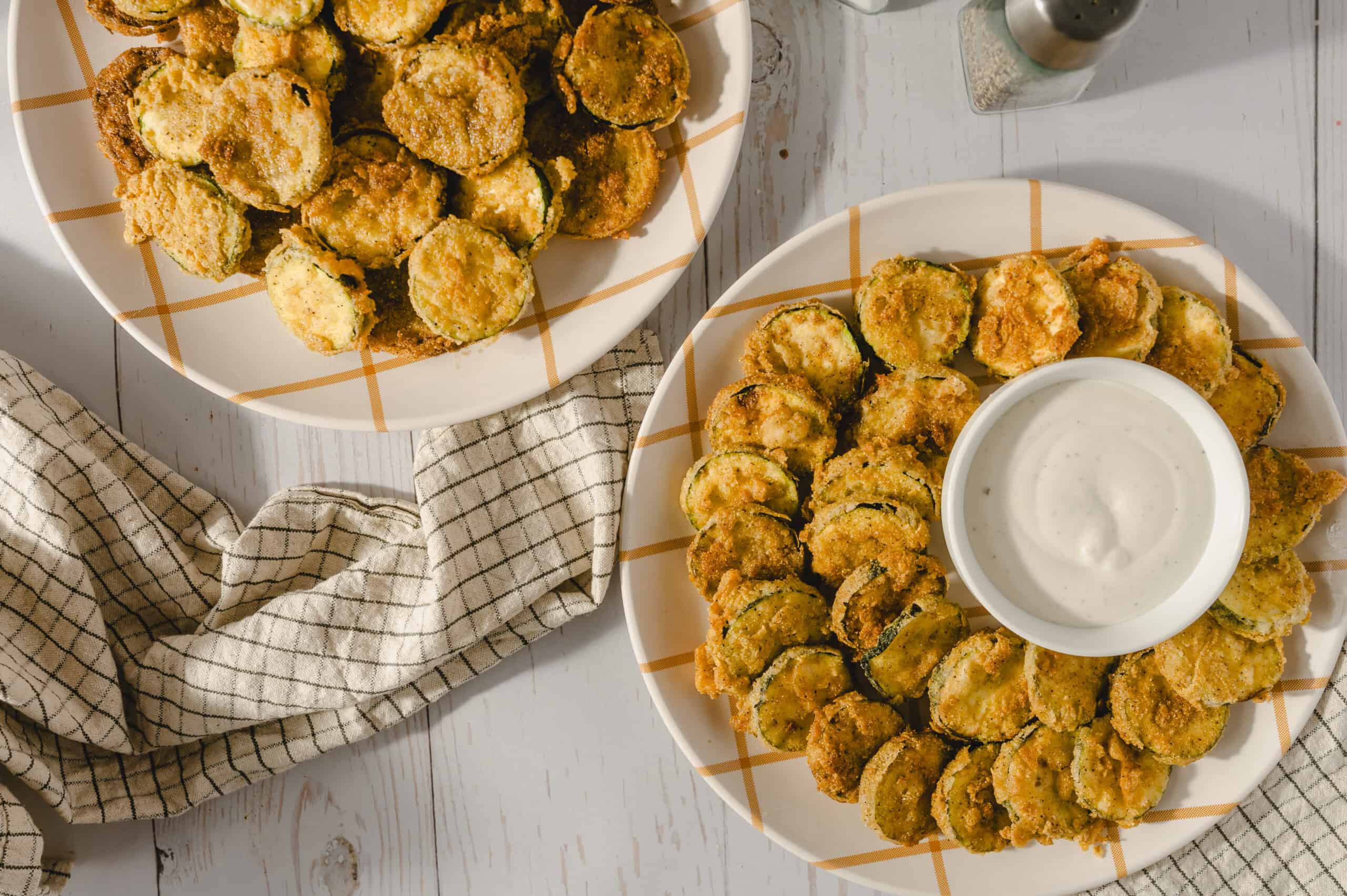 Crispy fried zucchini rounds served with creamy dipping sauce on a checkered plate, perfect for appetizer or snack.