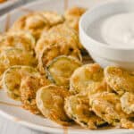 Golden fried zucchini chips with crispy breading served with a side of creamy dipping sauce on a white patterned plate. Perfect appetizer or snack, healthy zucchini recipe, vegetarian-friendly.