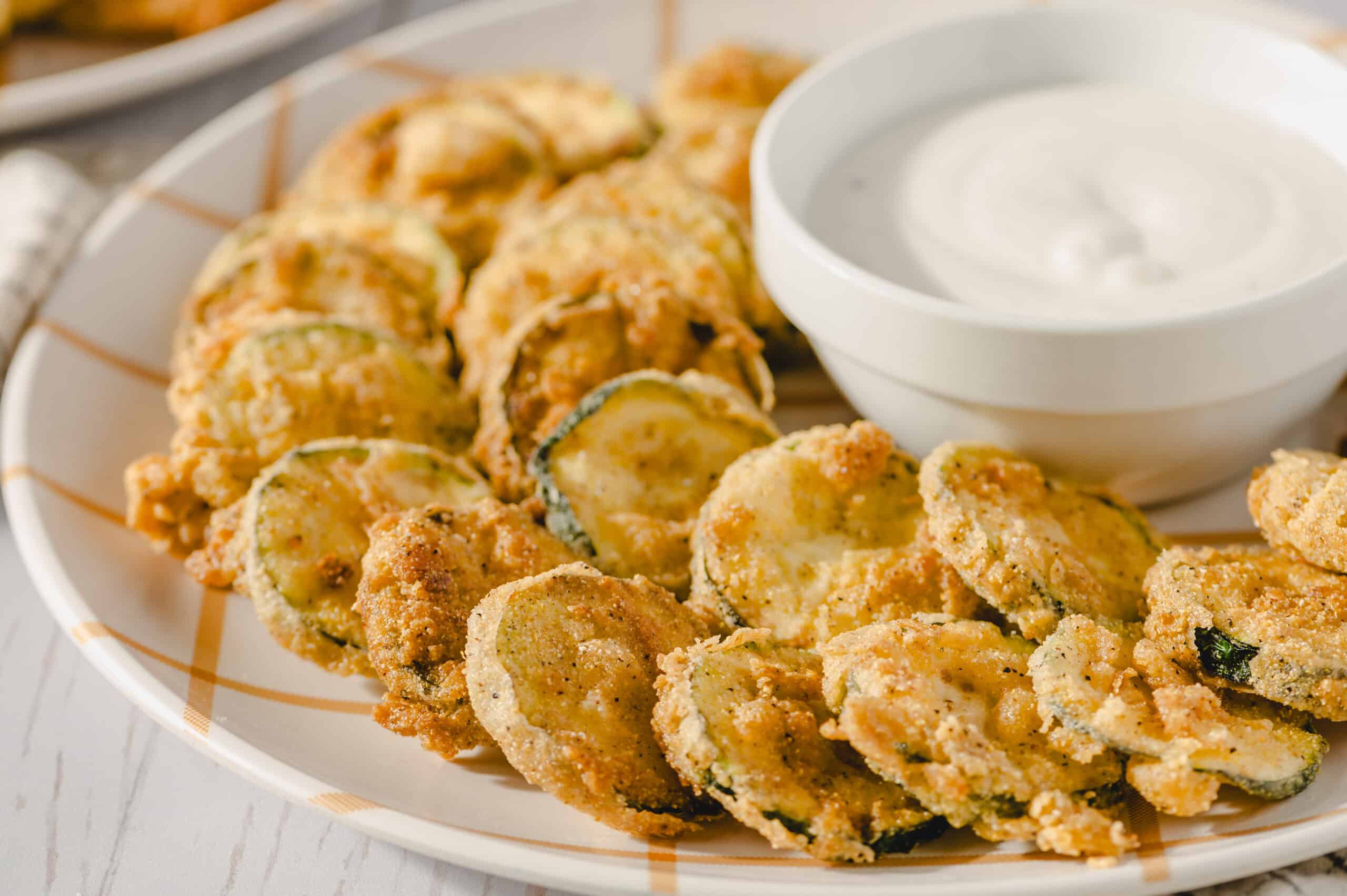 Golden fried zucchini chips with crispy breading served with a side of creamy dipping sauce on a white patterned plate. Perfect appetizer or snack, healthy zucchini recipe, vegetarian-friendly.