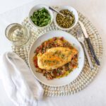 Juicy baked chicken breast served over vegetable medley with herbs, alongside fresh greens and lentils on a white plate, perfect for healthy meal ideas.