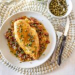 Roasted chicken breast with vegetable sauce on a white plate, garnished with herbs, served with a bowl of peppercorns and cutlery, ideal for healthy dinner recipes.