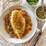 Golden baked chicken breast garnished with parsley, served over sautéed tomatoes and peppers, accompanied by fresh herbs and green peas, on a white plate for delicious healthy meal.