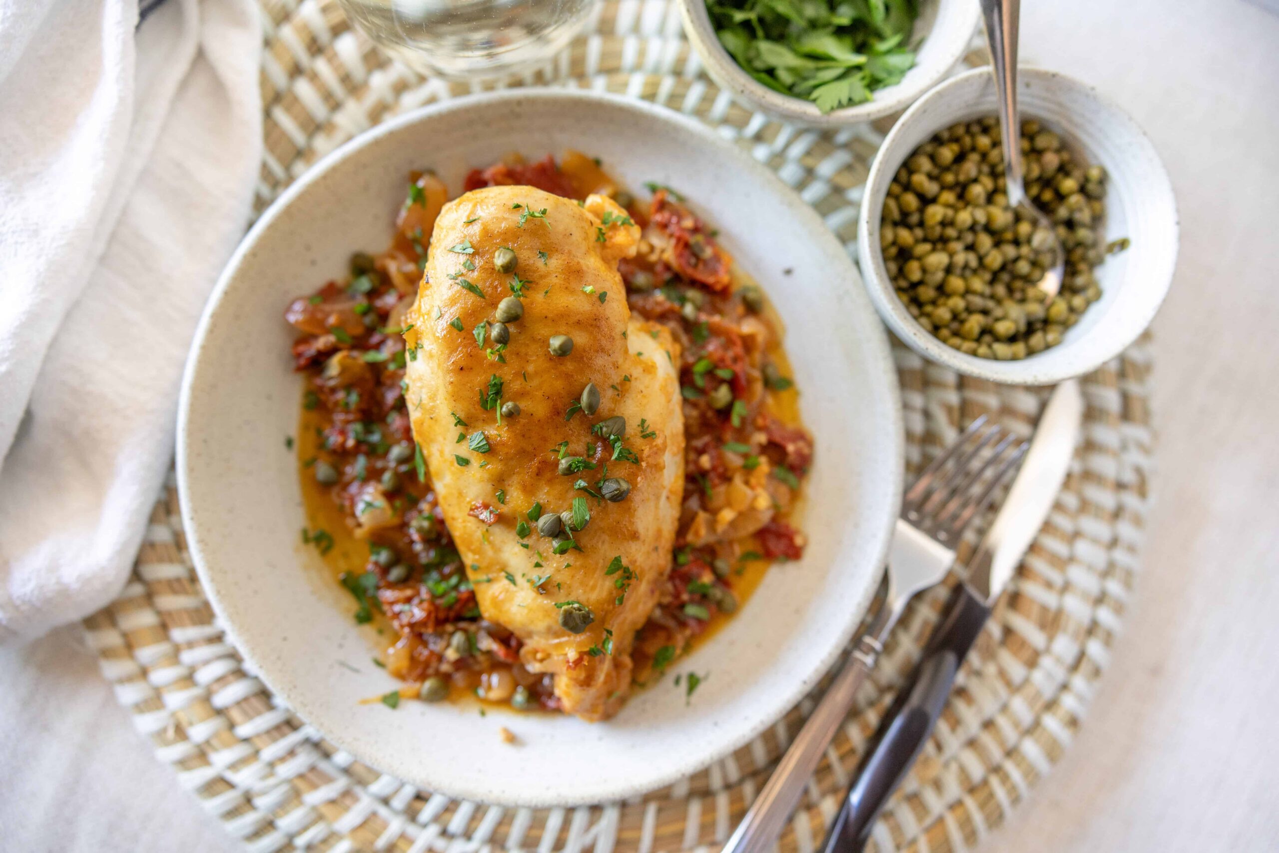 Golden baked chicken breast garnished with parsley, served over sautéed tomatoes and peppers, accompanied by fresh herbs and green peas, on a white plate for delicious healthy meal.
