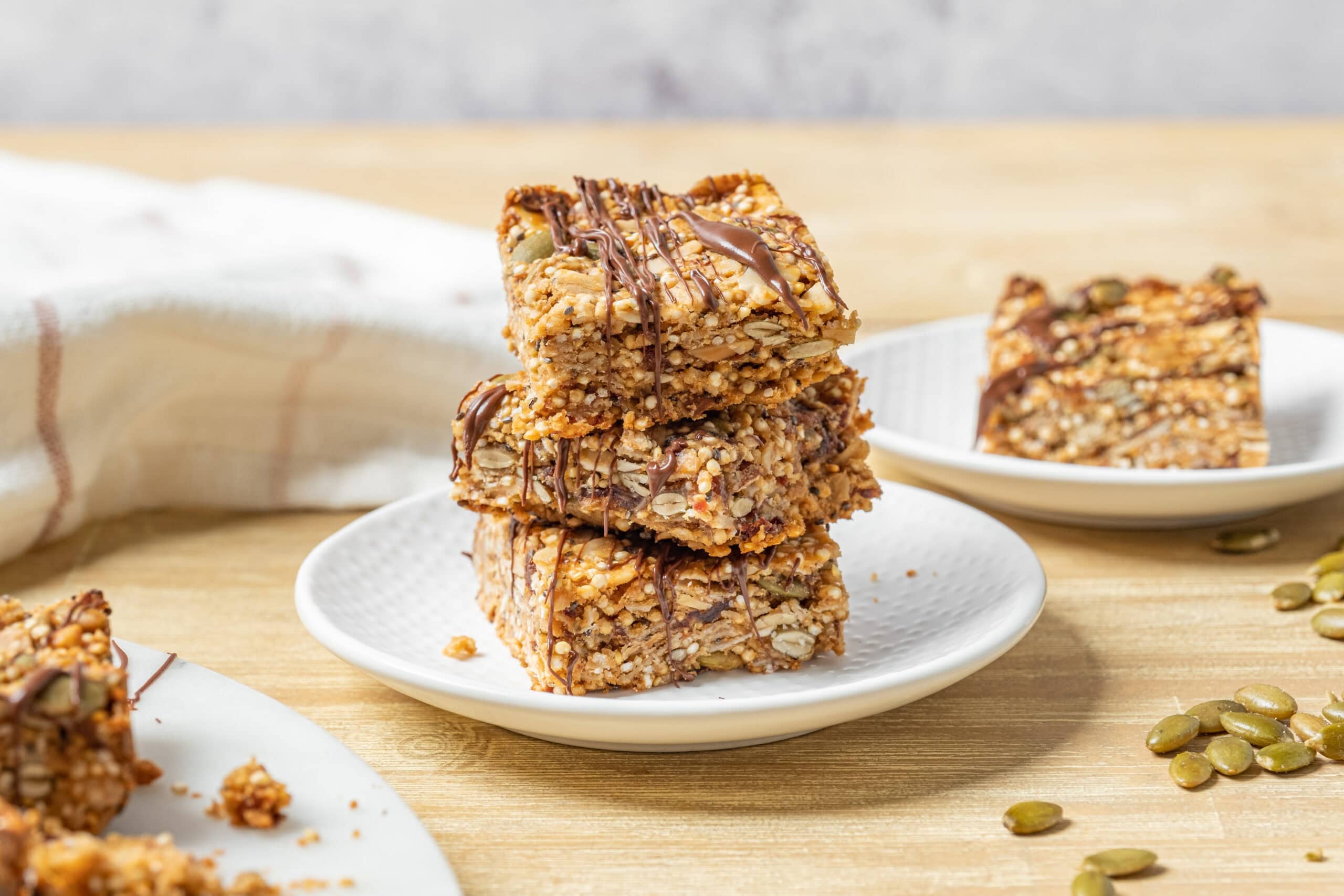 Crunchy homemade granola bar squares drizzled with chocolate on white plates, healthy snack with oats, seeds, and nuts. Perfect for energy, breakfast, or on-the-go treats, styled on a wooden table.
