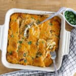 Creamy baked potato casserole with melted cheese and herbs in a white ceramic dish, served on a rustic wooden table with fresh chopped herbs on the side.