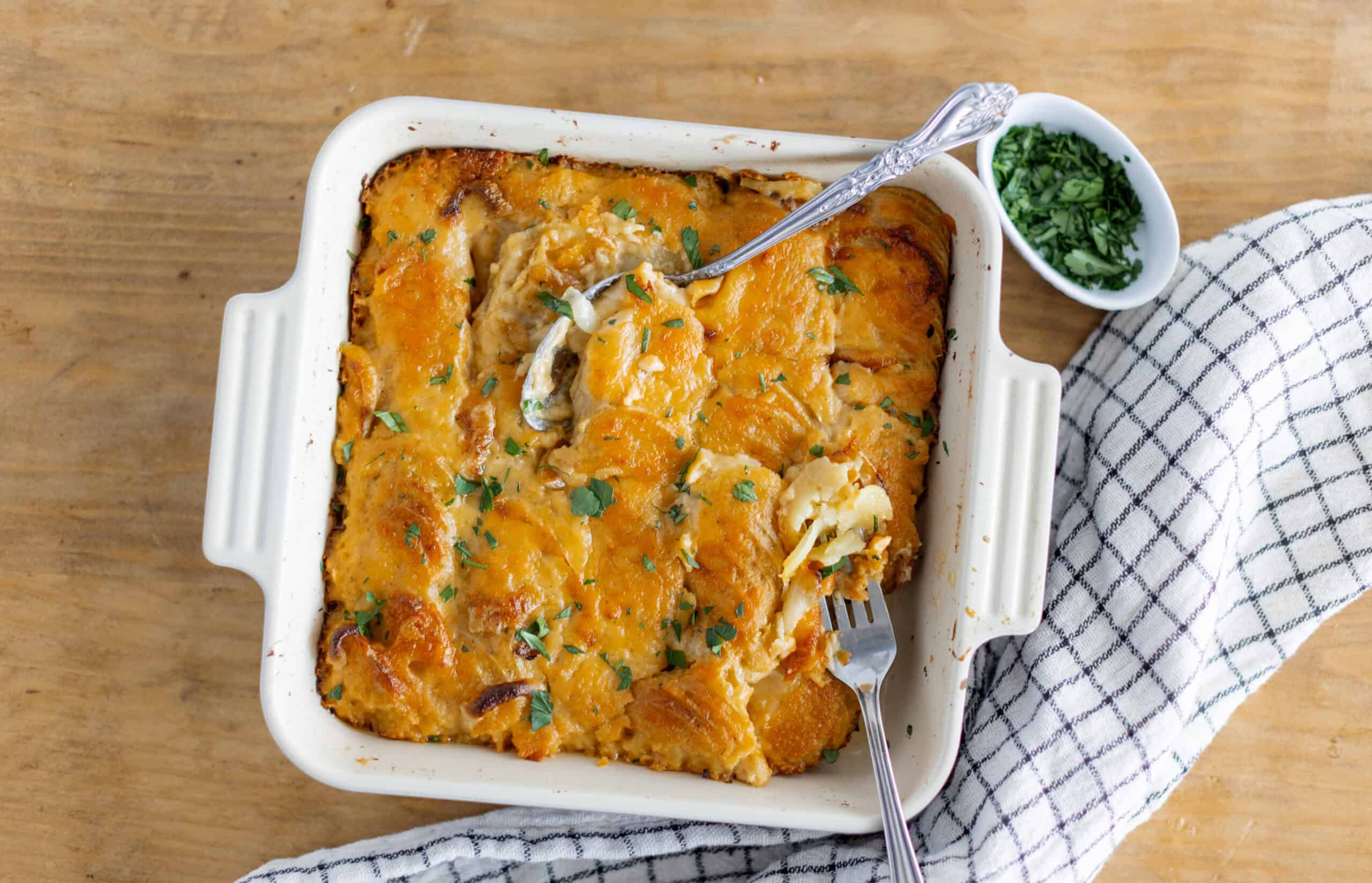 Creamy baked potato casserole with melted cheese and herbs in a white ceramic dish, served on a rustic wooden table with fresh chopped herbs on the side.