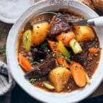 Hearty beef stew with potatoes, carrots, celery, and herbs in a rustic white bowl, served with bread on a wooden table, ideal for comfort food, slow cooking recipes, and family dinners.