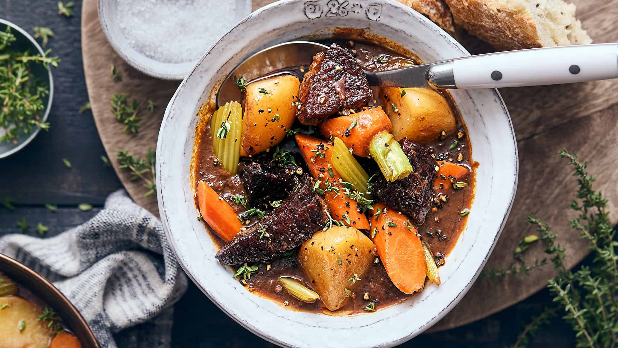 Hearty beef stew with potatoes, carrots, celery, and herbs in a rustic white bowl, served with bread on a wooden table, ideal for comfort food, slow cooking recipes, and family dinners.