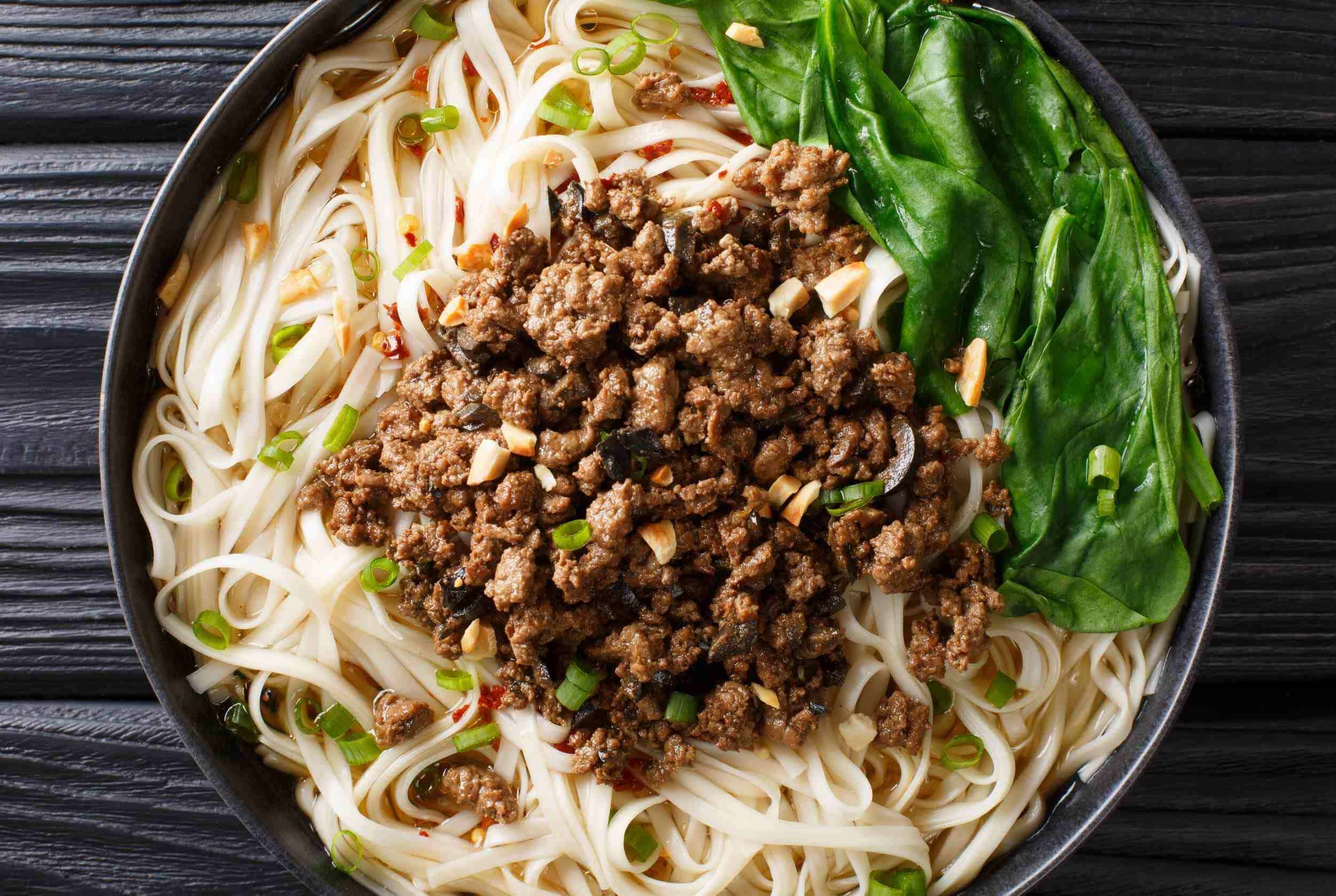 Savory Asian ground beef chow mein with noodles, fresh basil, and chopped green onions in a black bowl on rustic wooden table.