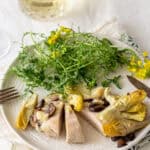 Fresh cooked chicken breast with artichokes and mushrooms served with a side of mixed greens and herbs on a white plate.