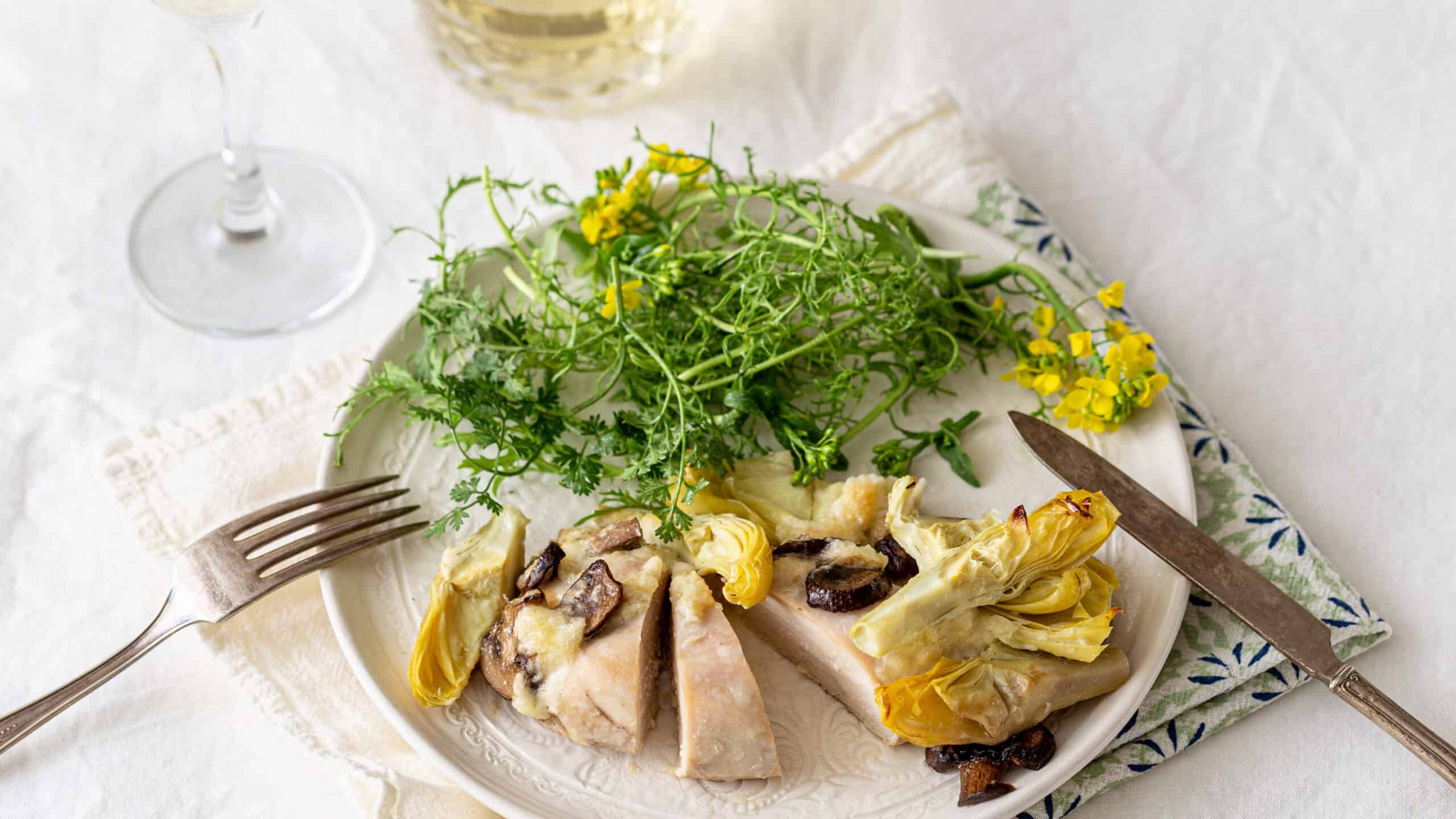 Fresh cooked chicken breast with artichokes and mushrooms served with a side of mixed greens and herbs on a white plate.