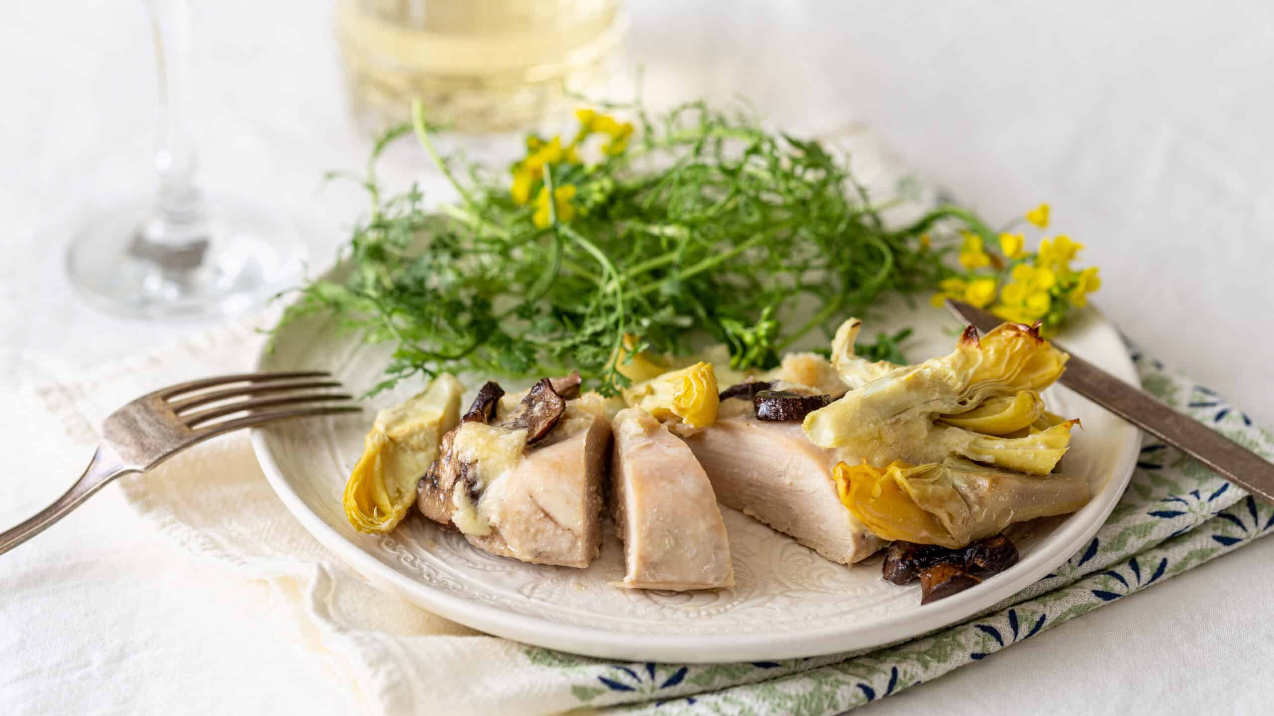 Succulent poached chicken breast topped with artichoke hearts and mushrooms, served with fresh greens and edible flowers on a white plate for a elegant and healthy meal.