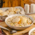 Creamy chicken and vegetable stew topped with steamed bread dumplings, served in a white bowl on a wooden cutting board. Freshly baked bread rolls are in a basket behind, with salt and pepper shakers nearby.