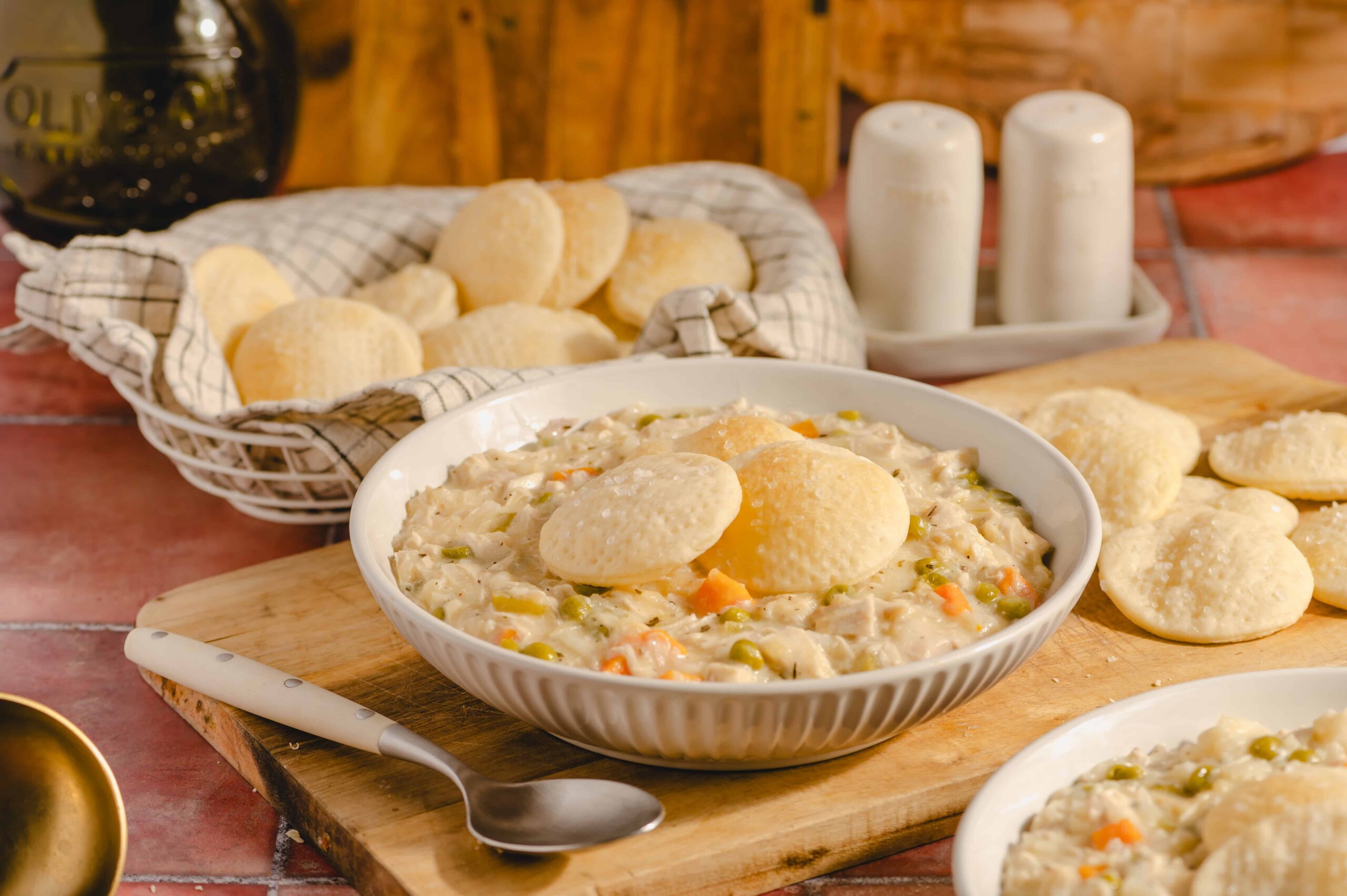 Creamy chicken and vegetable stew topped with steamed bread dumplings, served in a white bowl on a wooden cutting board. Freshly baked bread rolls are in a basket behind, with salt and pepper shakers nearby.