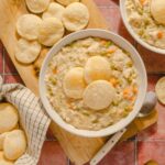 Creamy chicken pot pie soup garnished with melba toast in white bowls, surrounded by salt and pepper shakers, fresh melba toast on a wooden cutting board, and scattered melba toast on a rustic tiled countertop.