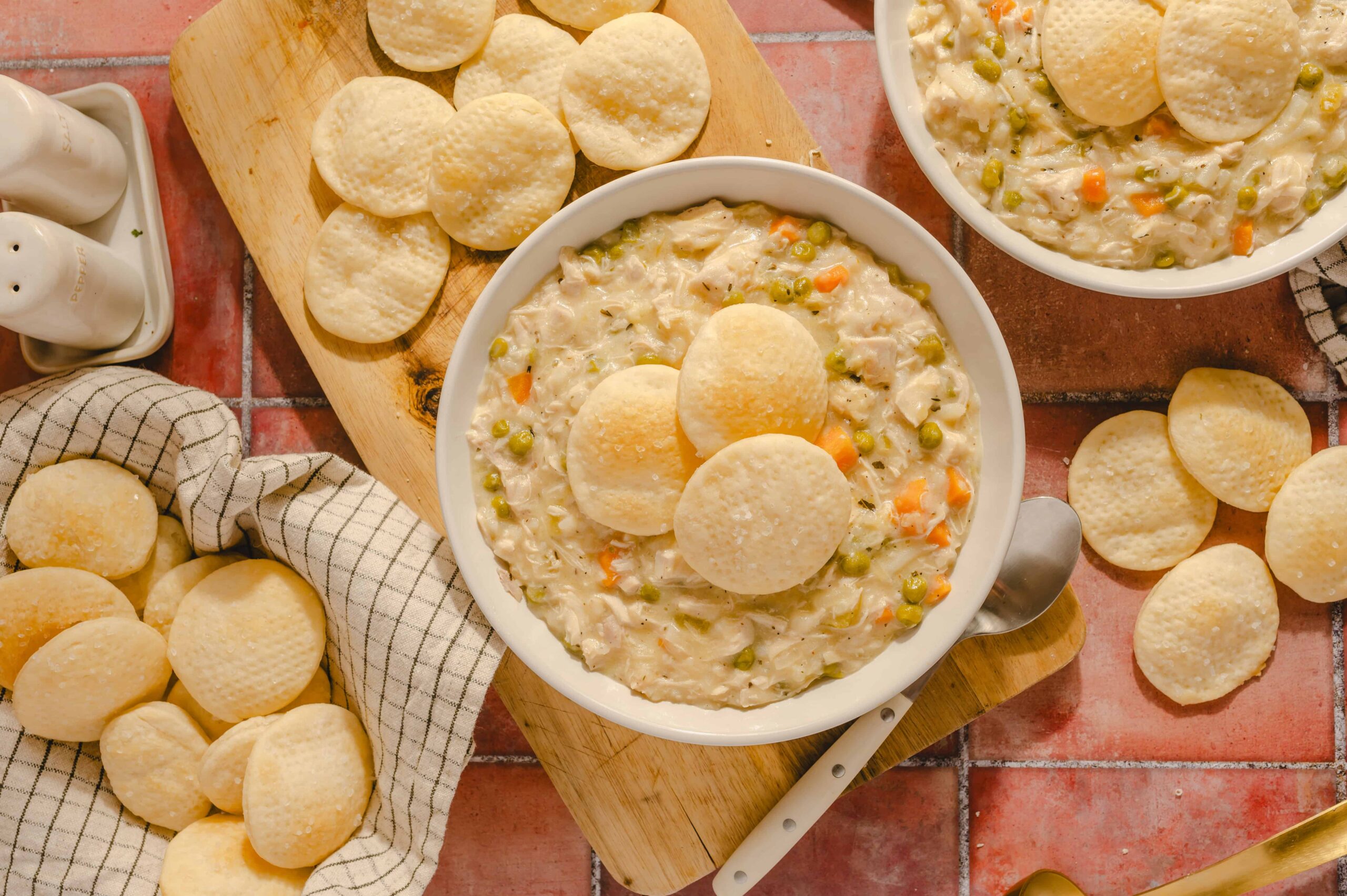 Creamy chicken pot pie soup garnished with melba toast in white bowls, surrounded by salt and pepper shakers, fresh melba toast on a wooden cutting board, and scattered melba toast on a rustic tiled countertop.