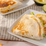 Delicious cheesy chicken quesadilla wrapped in a flour tortilla, served on a white plate with fresh avocado and shredded cheese nearby. Perfect for quick, flavorful Mexican-inspired meals.