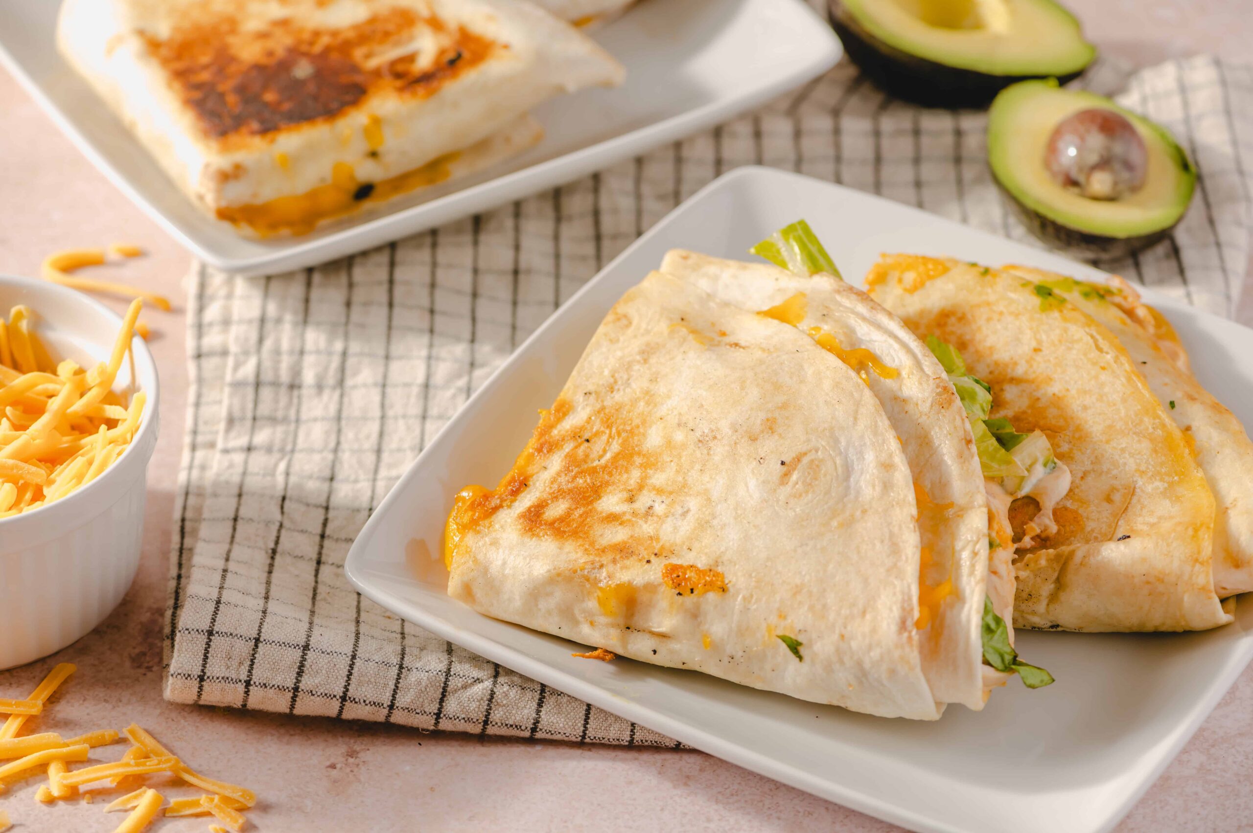 Delicious cheesy chicken quesadilla wrapped in a flour tortilla, served on a white plate with fresh avocado and shredded cheese nearby. Perfect for quick, flavorful Mexican-inspired meals.