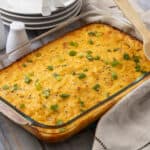 Creamy baked macaroni and cheese in a glass dish topped with chopped green onions, served on a rustic wooden table with plates and salt & pepper shakers in the background.