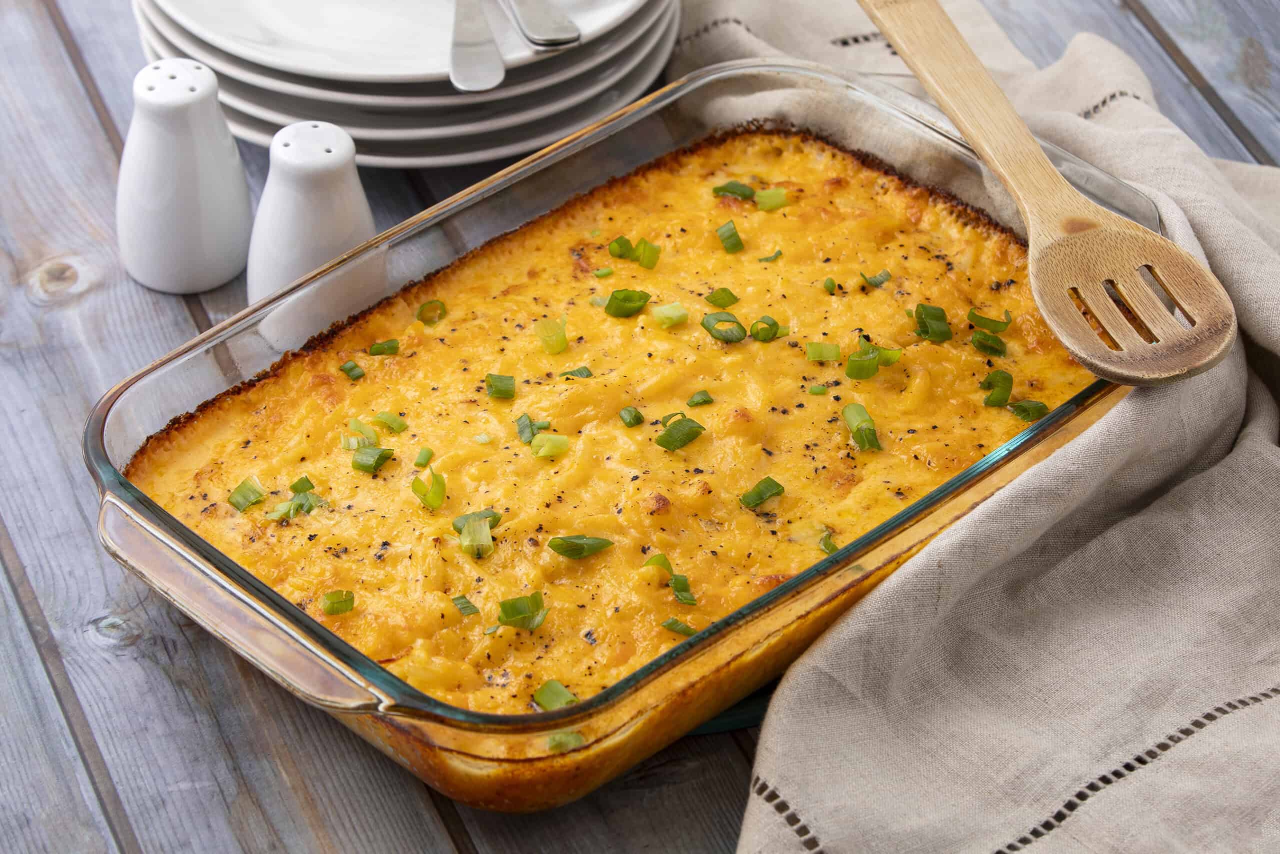 Creamy baked macaroni and cheese in a glass dish topped with chopped green onions, served on a rustic wooden table with plates and salt & pepper shakers in the background.