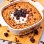 Creamy oatmeal casserole topped with fresh blackberries, whipped cream, and chocolate chips, served in a decorative ceramic dish on a wooden cutting board. Nutritious breakfast or dessert option.