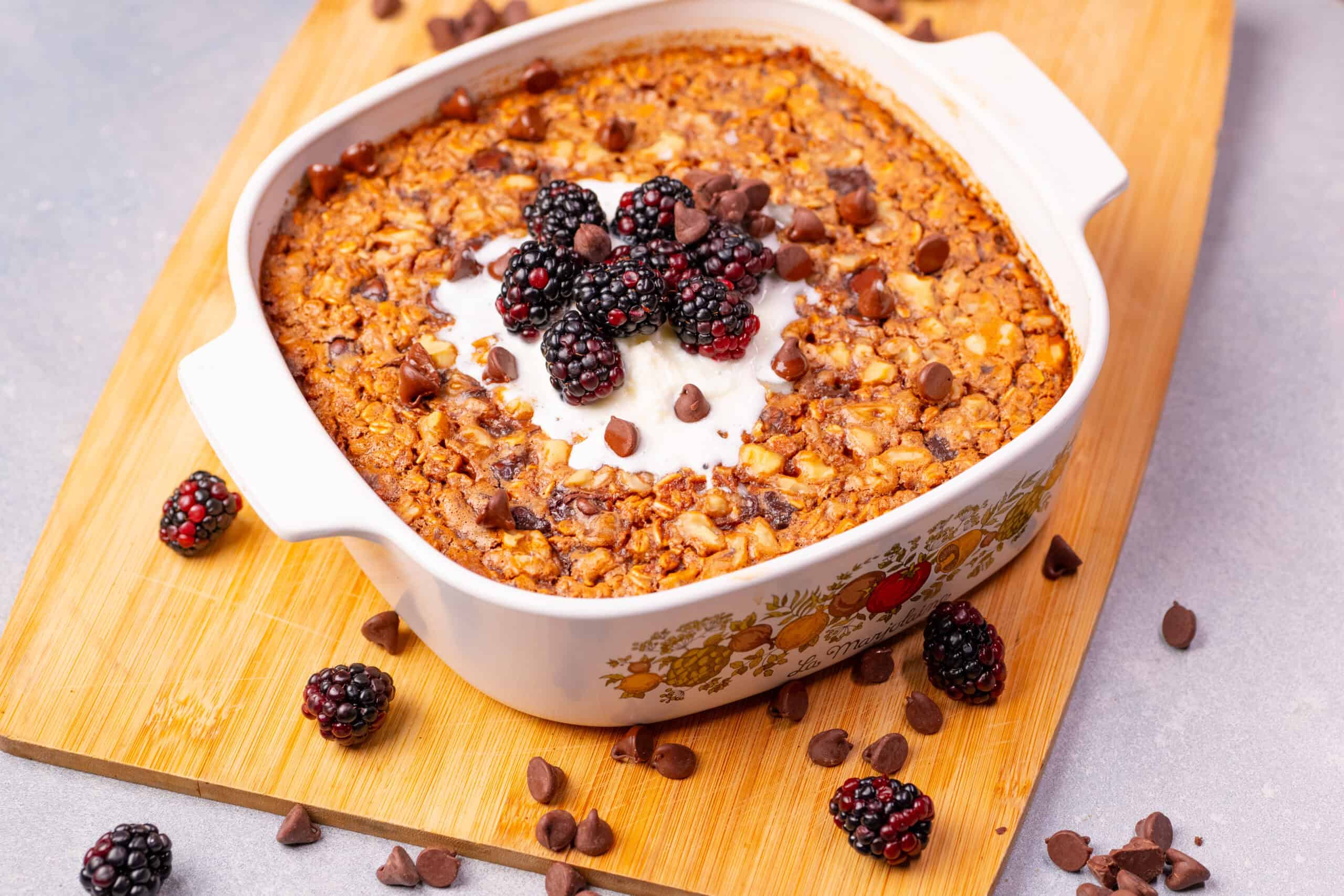 Creamy oatmeal casserole topped with fresh blackberries, whipped cream, and chocolate chips, served in a decorative ceramic dish on a wooden cutting board. Nutritious breakfast or dessert option.