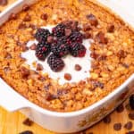 Rich dessert casserole with blackberries, chocolate chips, and creamy topping in a white baking dish on a wooden surface.