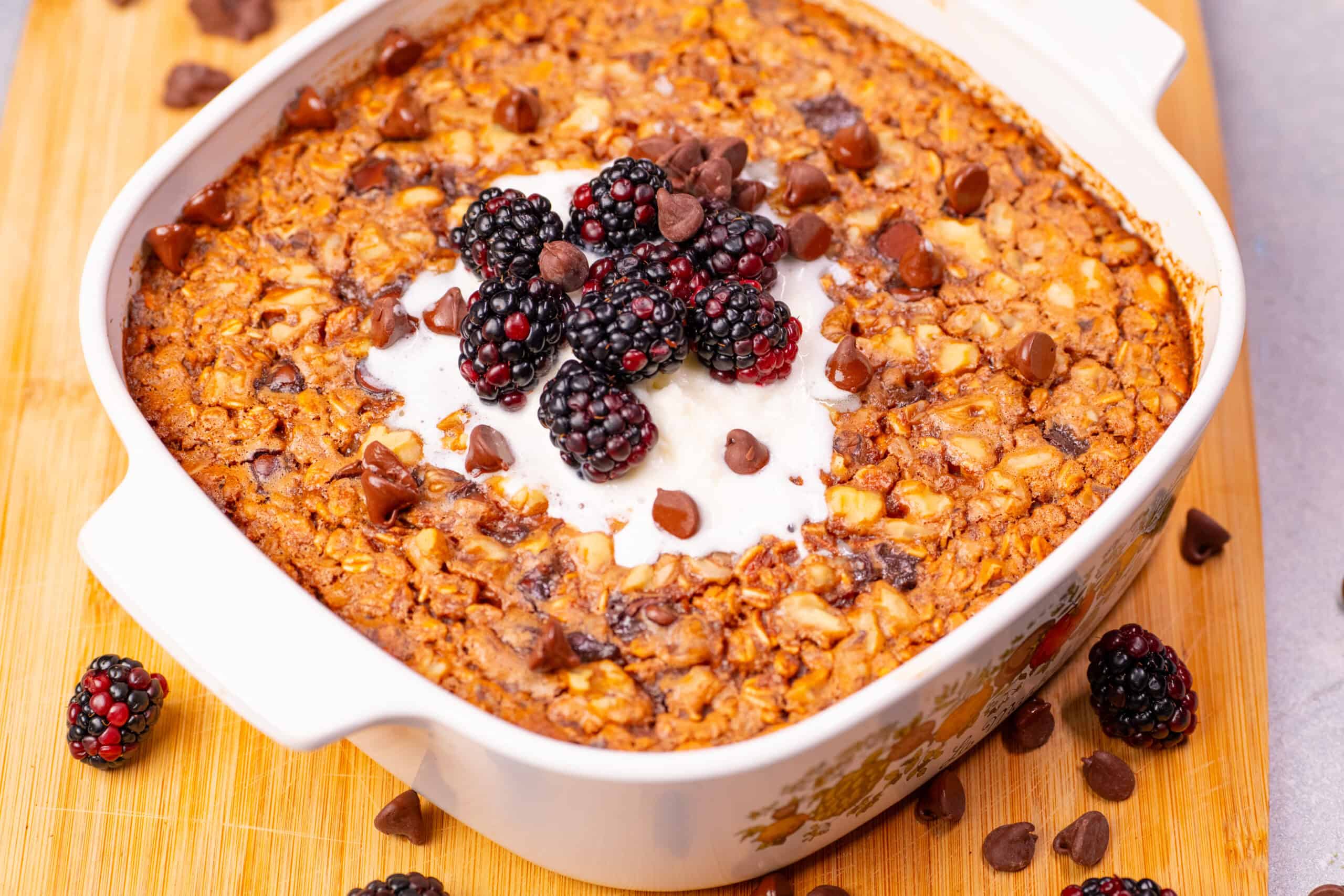 Rich dessert casserole with blackberries, chocolate chips, and creamy topping in a white baking dish on a wooden surface.
