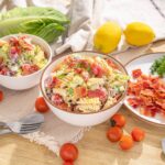 Fresh pasta salad with cherry tomatoes, bacon, lettuce, and dressing, served in bowls on a rustic wooden table with lemon, cherry tomatoes, lettuce, and herbs for a delicious summer meal.