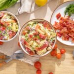 Fresh pasta salad with cherry tomatoes, crispy bacon, and leafy greens on a rustic wooden table, garnished with dill, perfect for summer or casual meals.