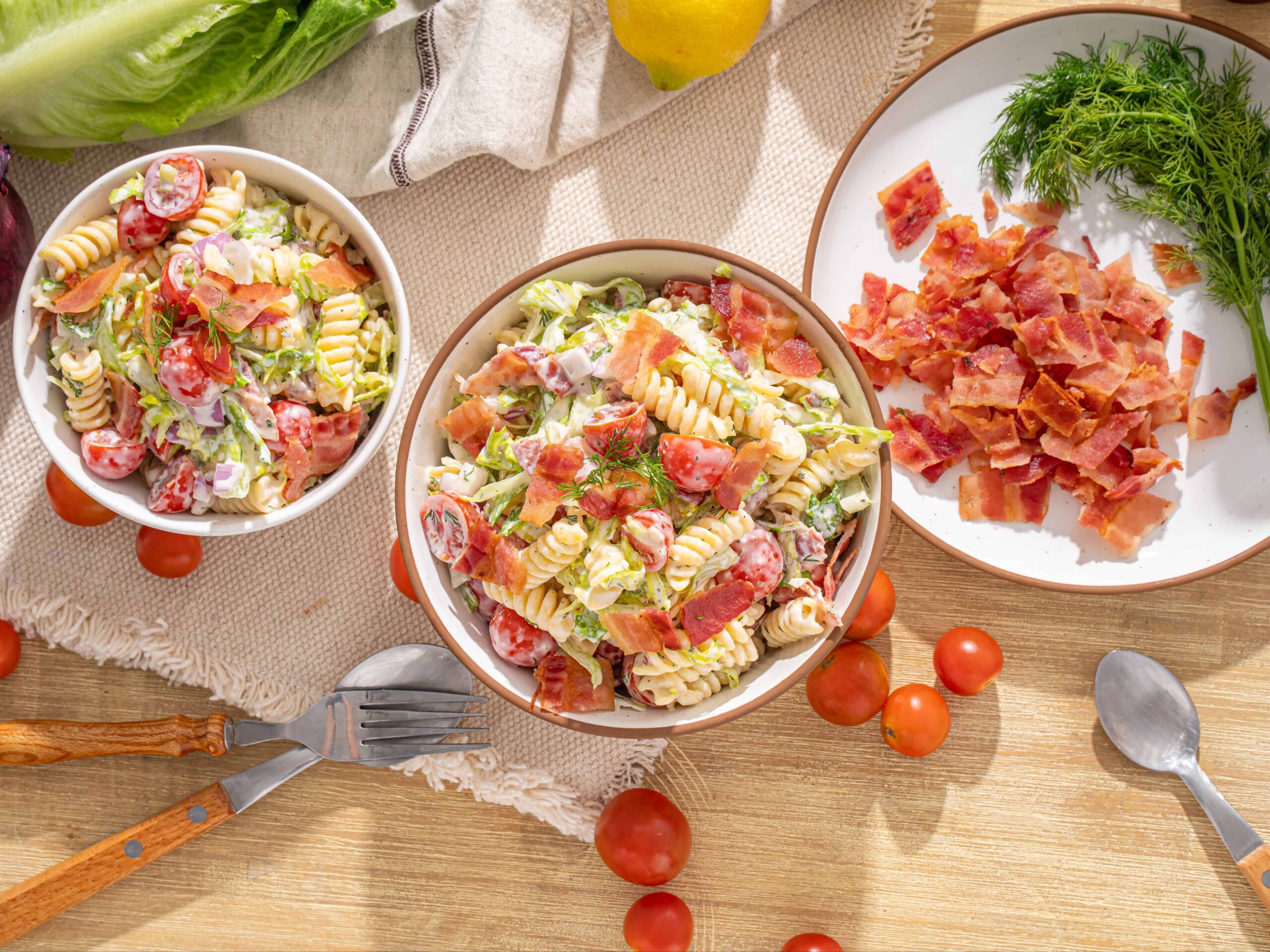 Fresh pasta salad with cherry tomatoes, crispy bacon, and leafy greens on a rustic wooden table, garnished with dill, perfect for summer or casual meals.