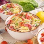 Creamy pasta salad with cherry tomatoes, bacon, and fresh vegetables in a white bowl, served on a rustic table with lemons, cherry tomatoes, and fresh greens for a fresh, flavorful summer dish.