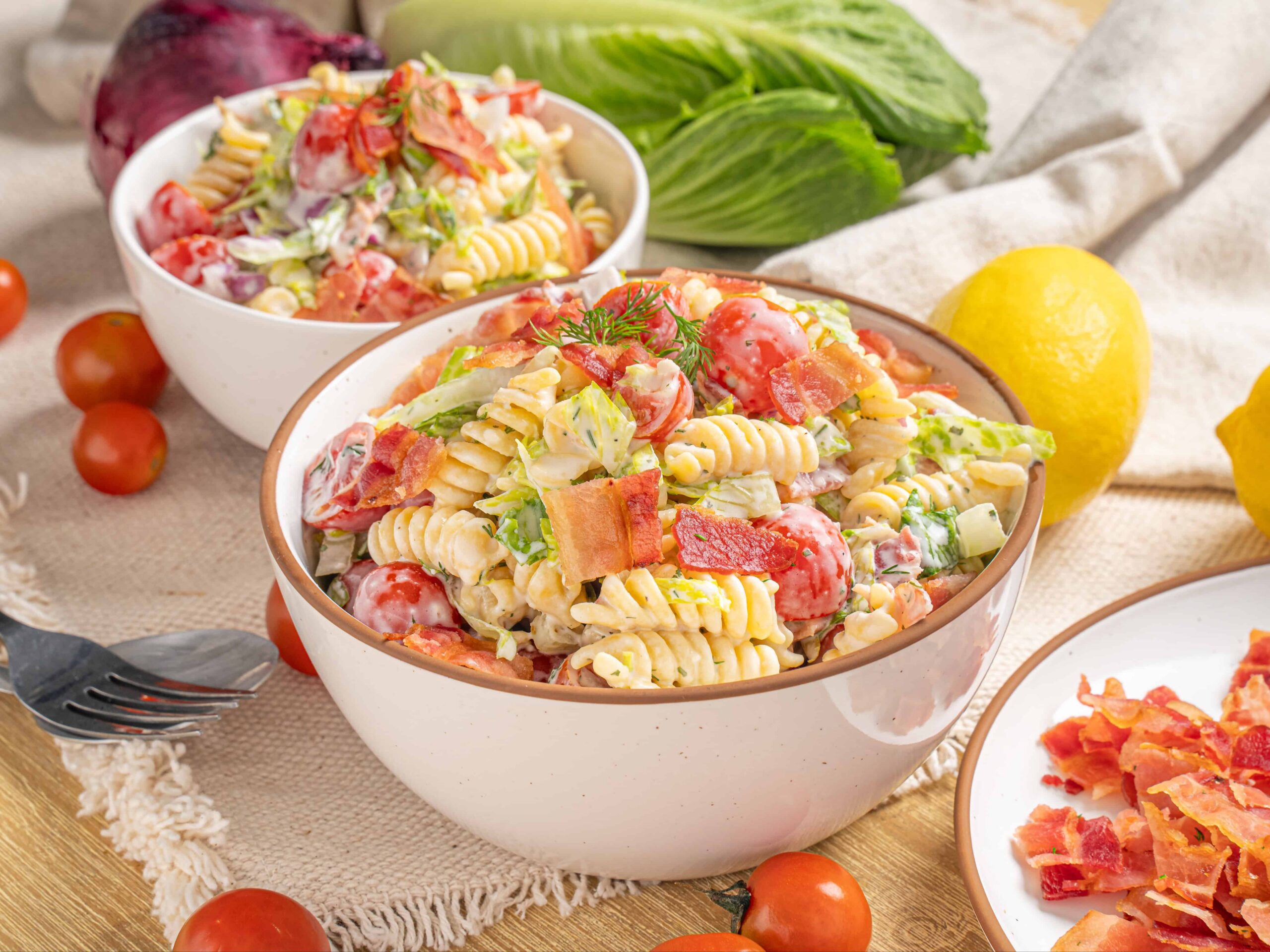 Creamy pasta salad with cherry tomatoes, bacon, and fresh vegetables in a white bowl, served on a rustic table with lemons, cherry tomatoes, and fresh greens for a fresh, flavorful summer dish.