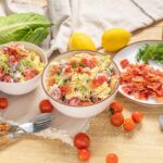 Creamy pasta salad with fresh cherry tomatoes, lettuce, bacon bits, and herbs on a rustic wooden table. Perfect for summer picnics and gatherings with vibrant ingredients and homemade flavor.