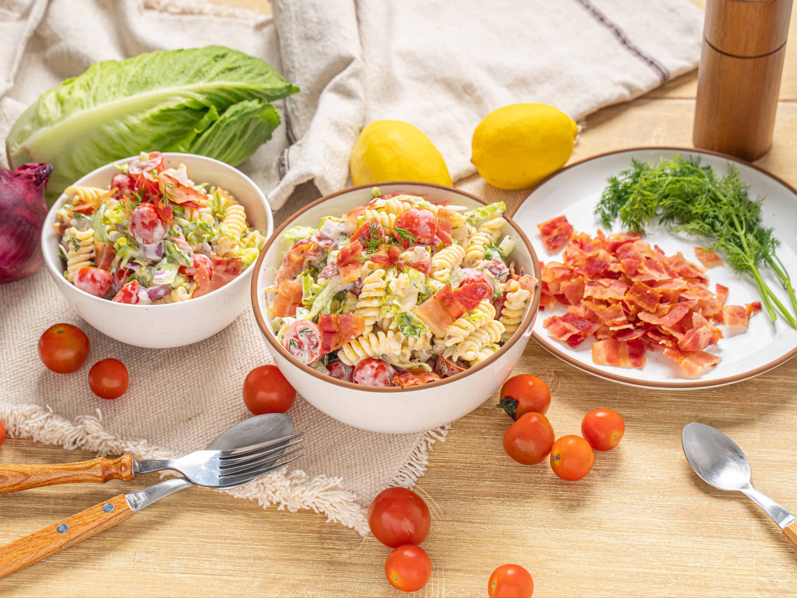 Creamy pasta salad with fresh cherry tomatoes, lettuce, bacon bits, and herbs on a rustic wooden table. Perfect for summer picnics and gatherings with vibrant ingredients and homemade flavor.
