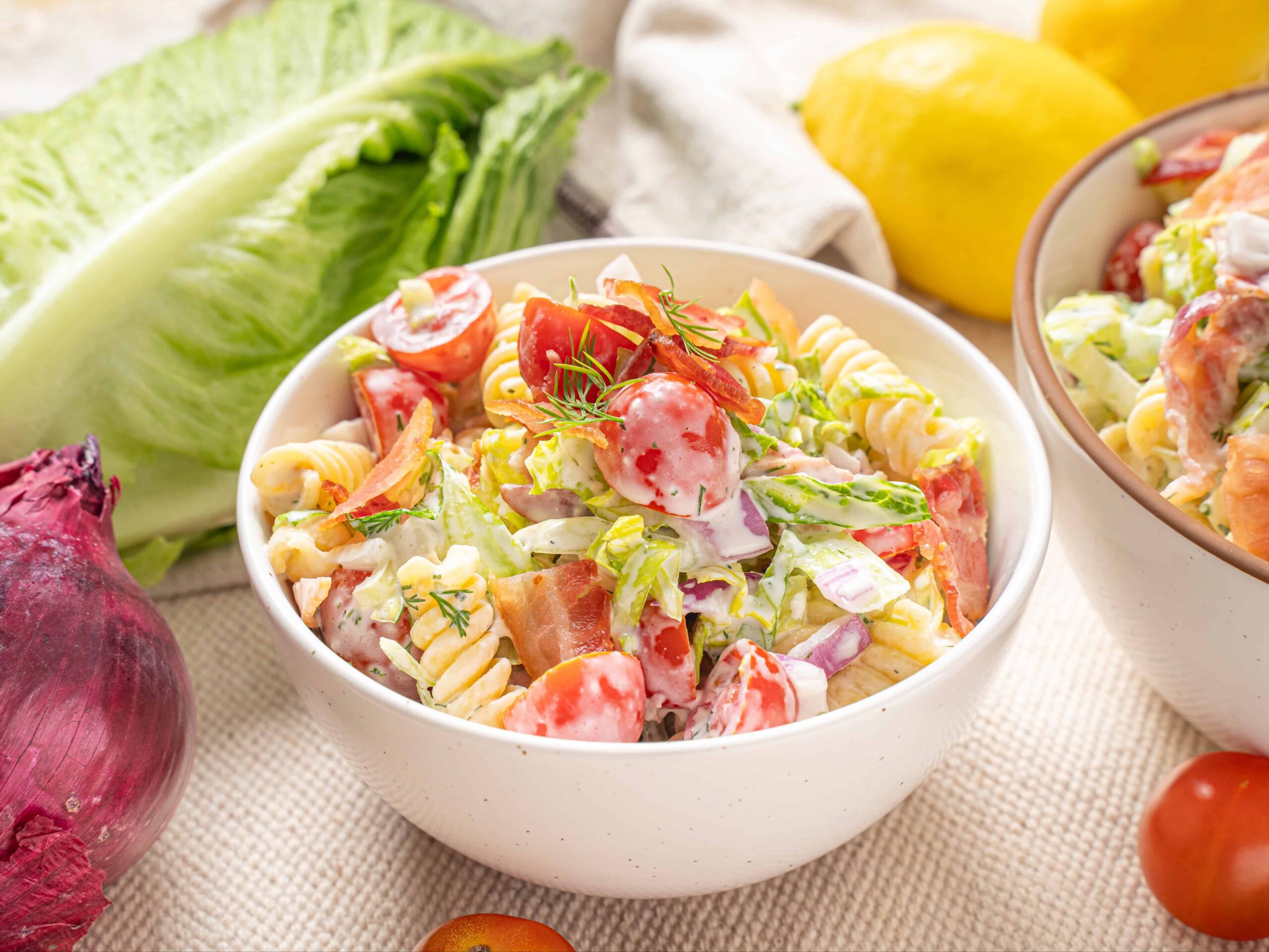 Fresh homemade pasta salad with cherry tomatoes, lettuce, cucumbers, and a creamy dressing in a white bowl, surrounded by raw vegetables and fresh ingredients for healthy recipes.