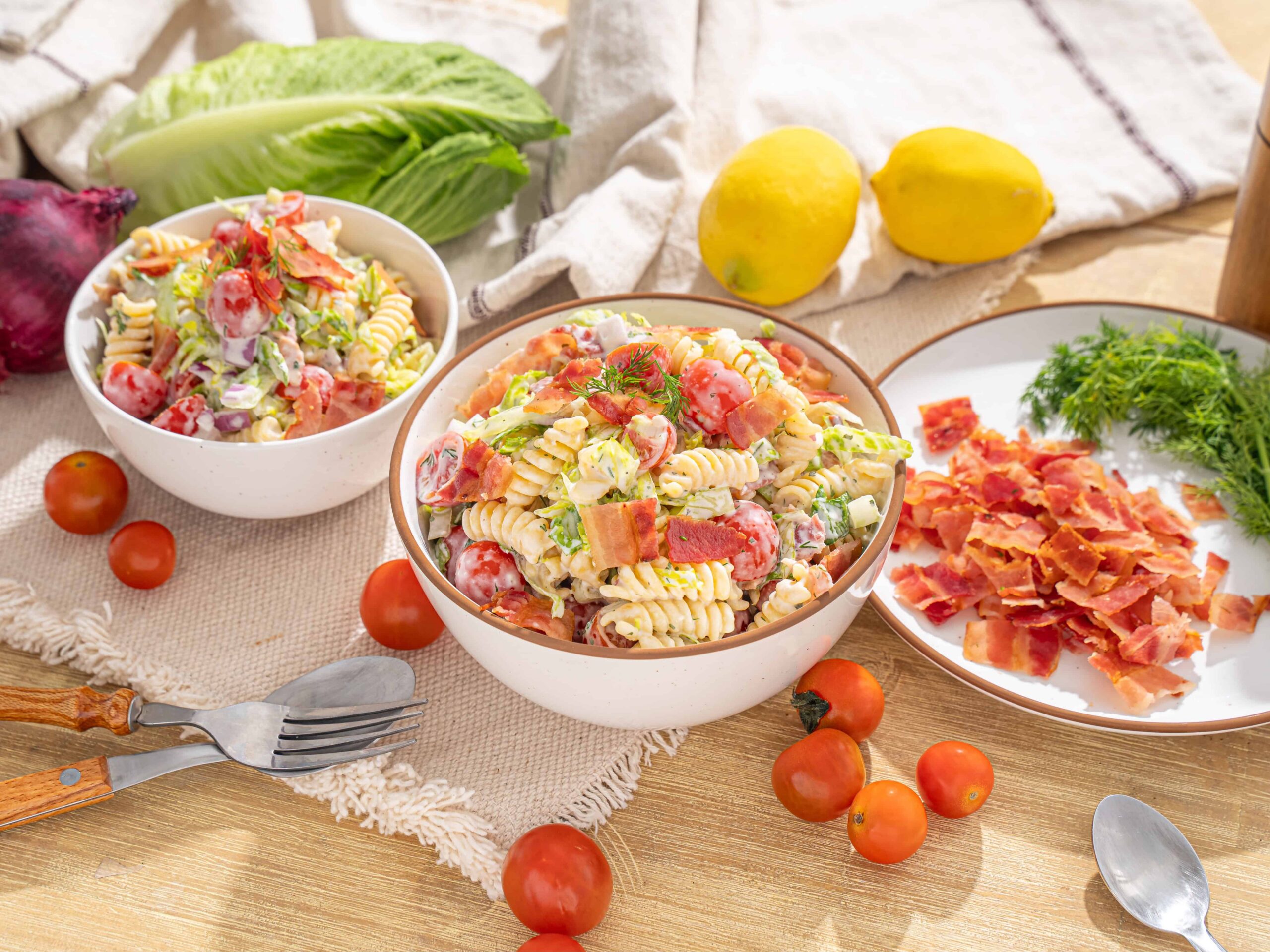 Fresh pasta salad with cherry tomatoes, bacon, lettuce, and dressing, served in bowls on a rustic wooden table with lemon, cherry tomatoes, lettuce, and herbs for a delicious summer meal.