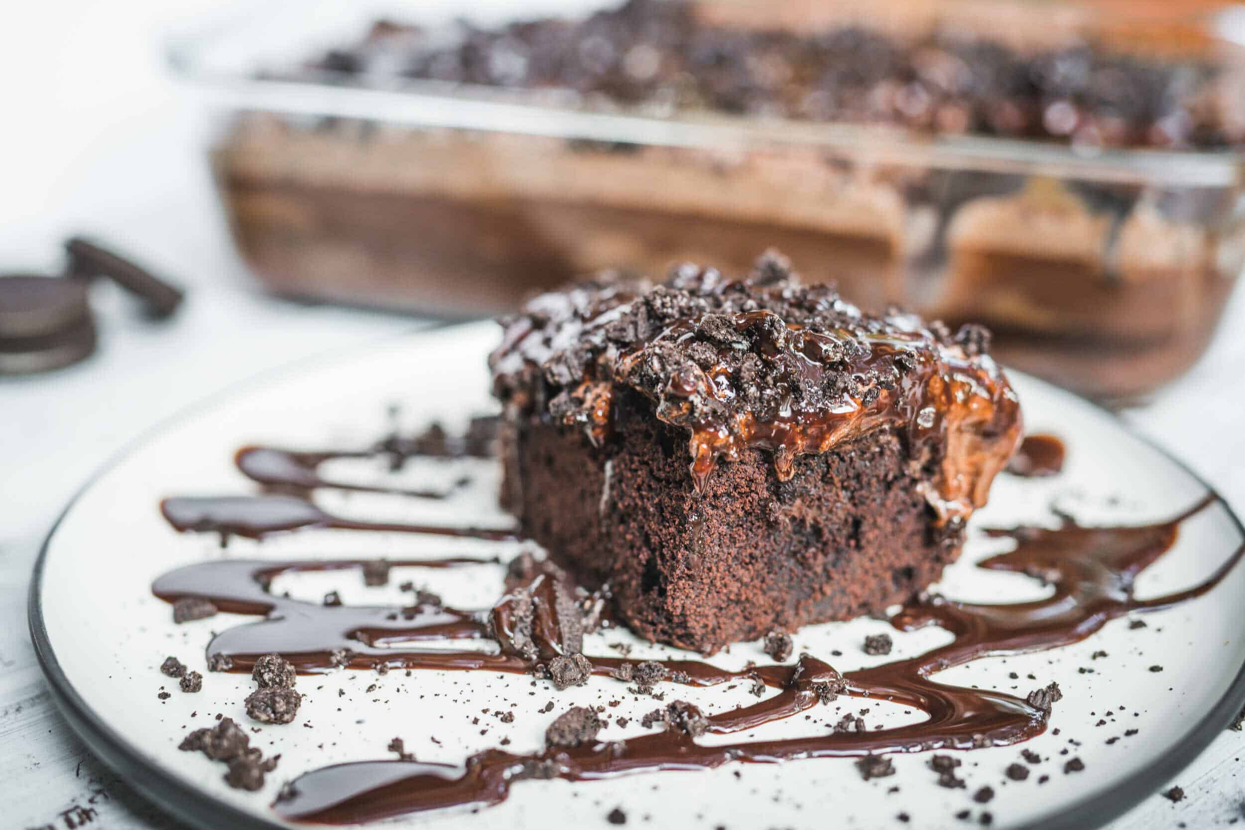Rich chocolate cake slice topped with chocolate ganache and cookie crumbles on a white plate with chocolate drizzle, perfect for dessert or special occasions.