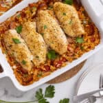 Baked chicken thighs on a bed of seasoned rice with vegetables and sausage in a white baking dish garnished with fresh parsley, served with white plates, forks, and a colorful patterned cloth.