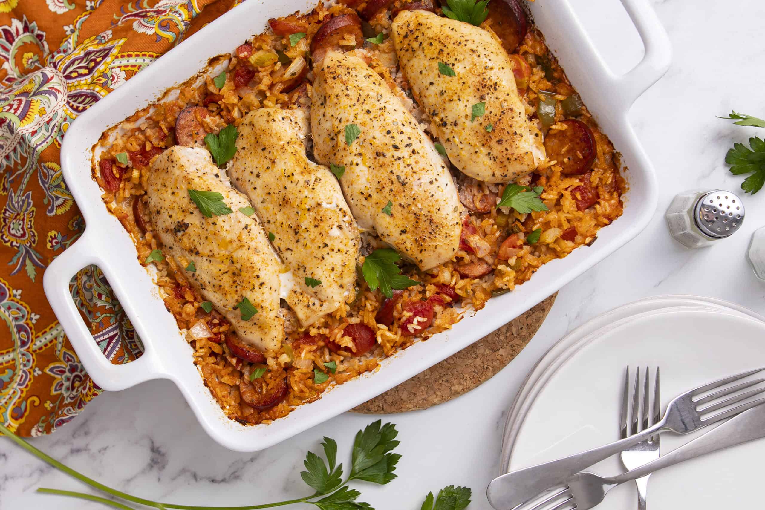 Baked chicken thighs on a bed of seasoned rice with vegetables and sausage in a white baking dish garnished with fresh parsley, served with white plates, forks, and a colorful patterned cloth.
