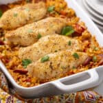 Juicy baked chicken breasts with rice and vegetables in a white baking dish. Perfect for a hearty, flavorful dinner recipe. Garnished with fresh parsley, served with stacked white plates.